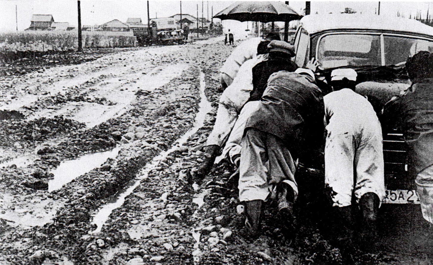 1950年代に日本で撮影された道路の写真。未舗装道路で動けなくなった自動車を複数の人が押している。出典：資料[2]