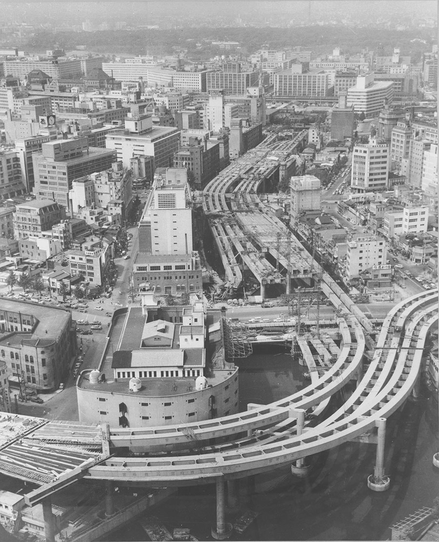 上記写真とほぼ同じアングルで撮影された、建設中の首都高速道路1号線（江戸橋ジャンクション付近）。中央付近が日本橋で、手前に佇むカーブを描く特徴的な建物は旧江戸橋倉庫ビル（現三菱倉庫江戸橋本館）だ。　写真提供：東京都（1963年11月1日撮影）